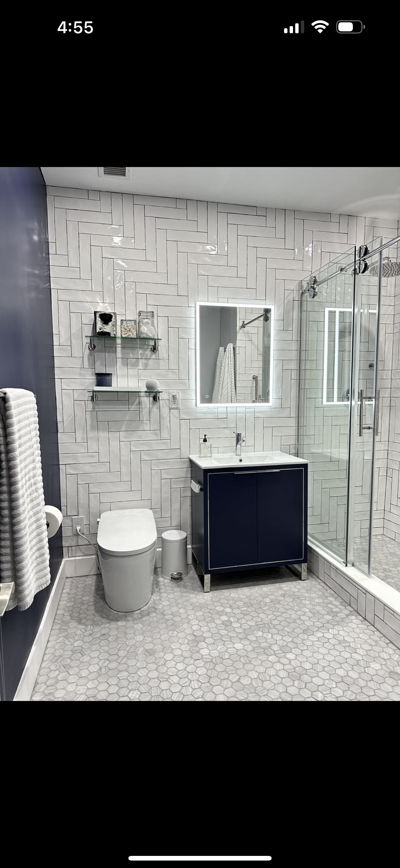 Modern bathroom with white and navy blue decor, featuring a toilet, a vanity with sink, a mirror with LED lighting, a glass shower enclosure, and decorative wall shelves.