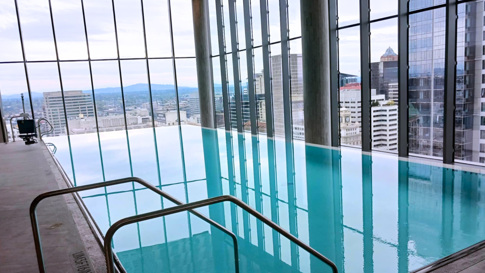 Indoor rooftop swimming pool with large glass windows overlooking a city skyline and mountains in the distance.