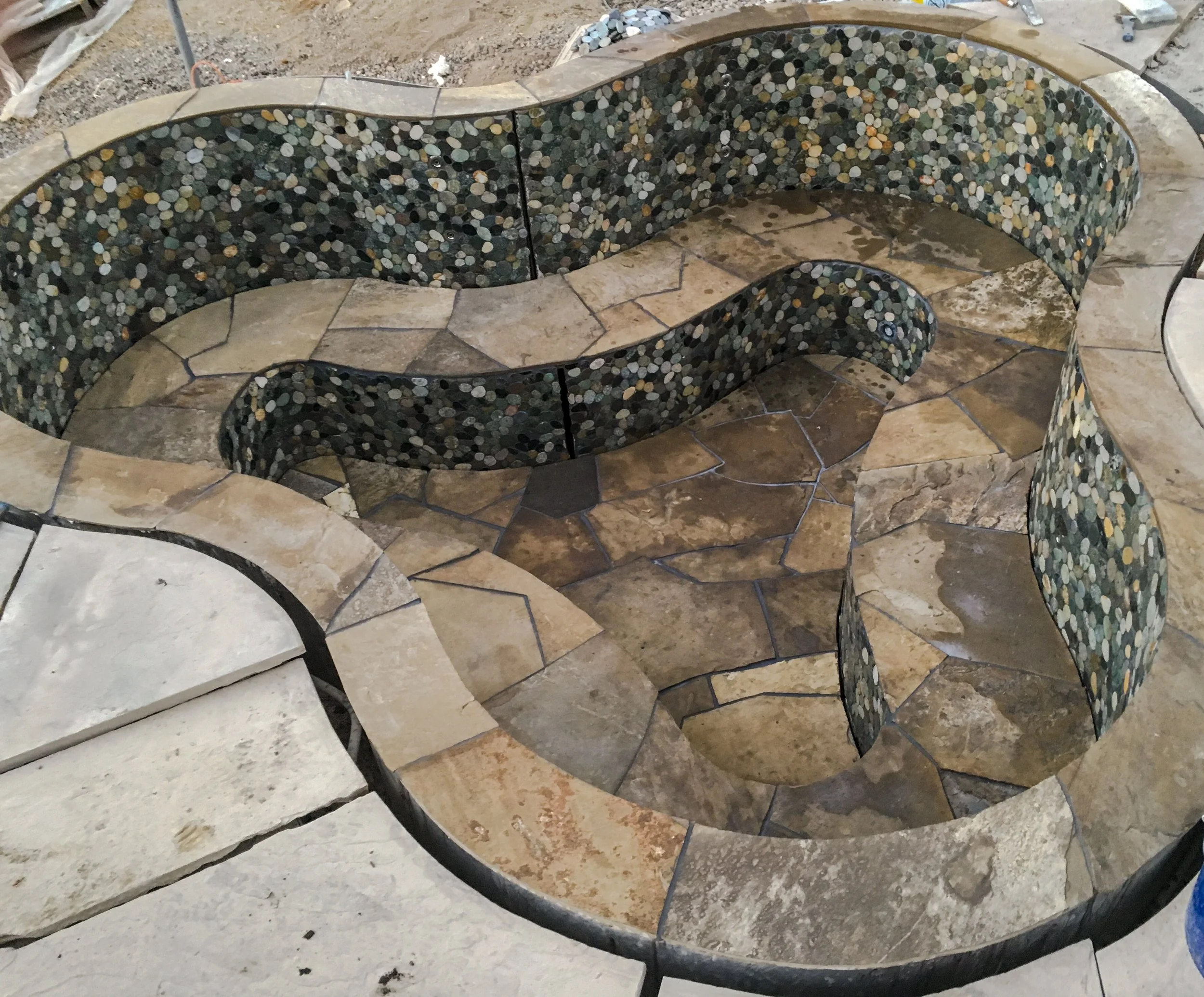 A custom stone bench with a curvy shape, featuring a mosaic pattern of small, multicolored stones on the backrest and armrest, and a stone tile seat and base.