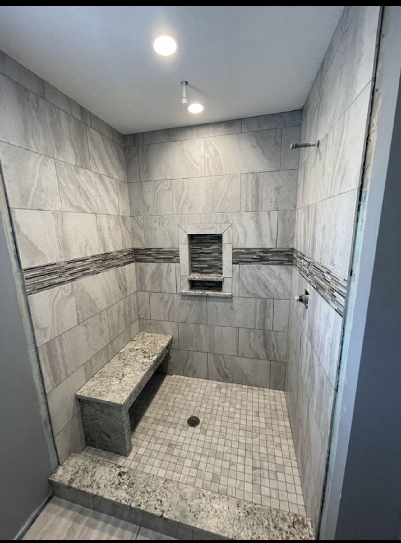 Shower with beige and gray stone tile walls, a built-in niche with decorative tile, small granite bench, and a tiled floor with a drain.