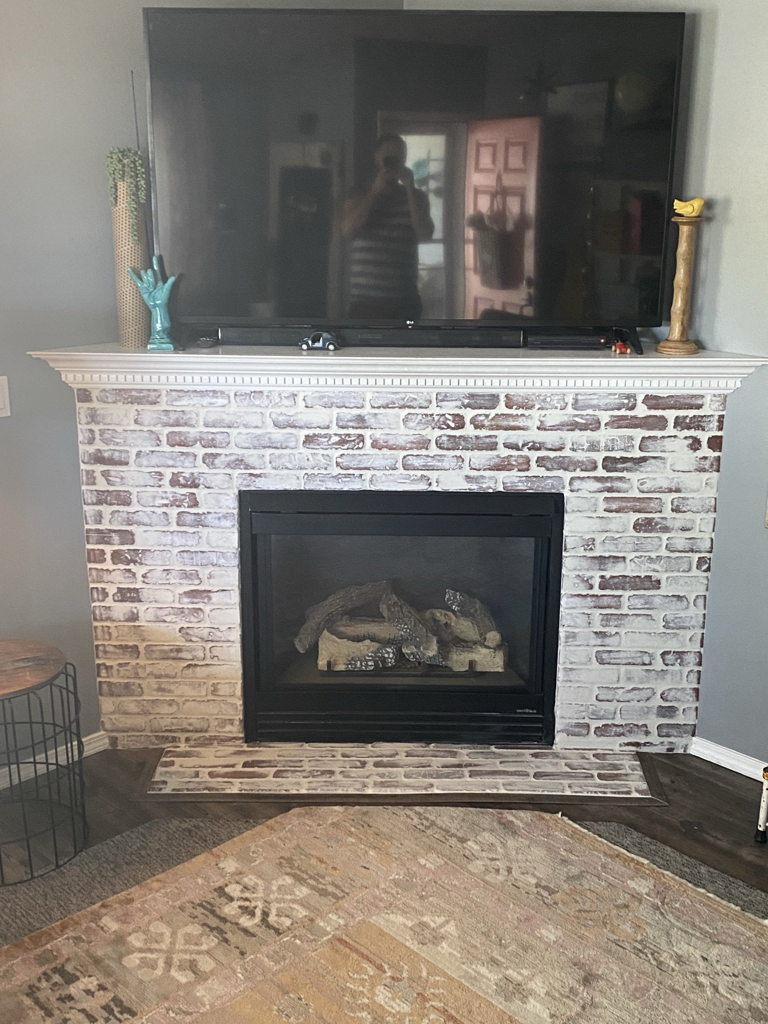 Living room fireplace with a large flat-screen TV on the mantle, which has decorative items including a green glove sculpture and a tall candlestick. The fireplace has a brick surround and inside there are decorative logs.