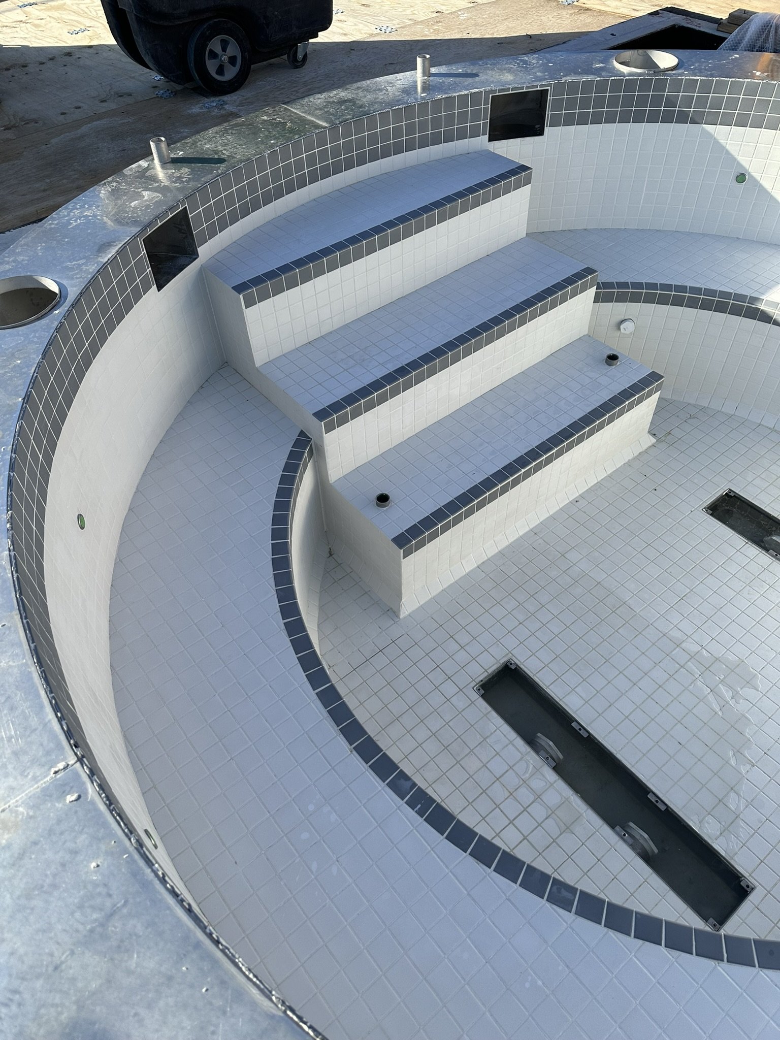 Empty circular hot tub with white and dark gray tiles, showing steps inside and black plumbing fittings, set on a project site.