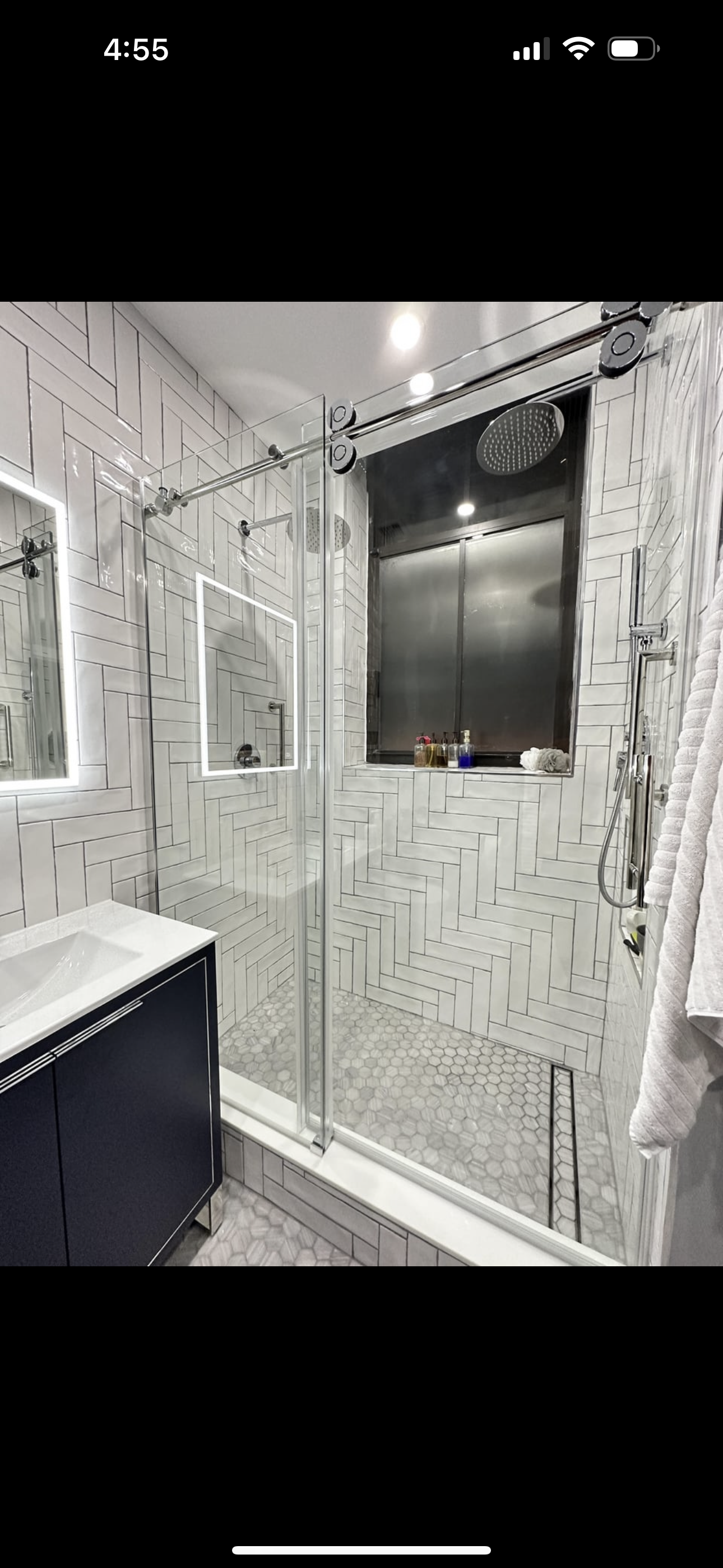 Modern bathroom with a walk-in glass shower featuring white herringbone tiles, hexagonal floor tiles, a black vanity, and a large frosted window.