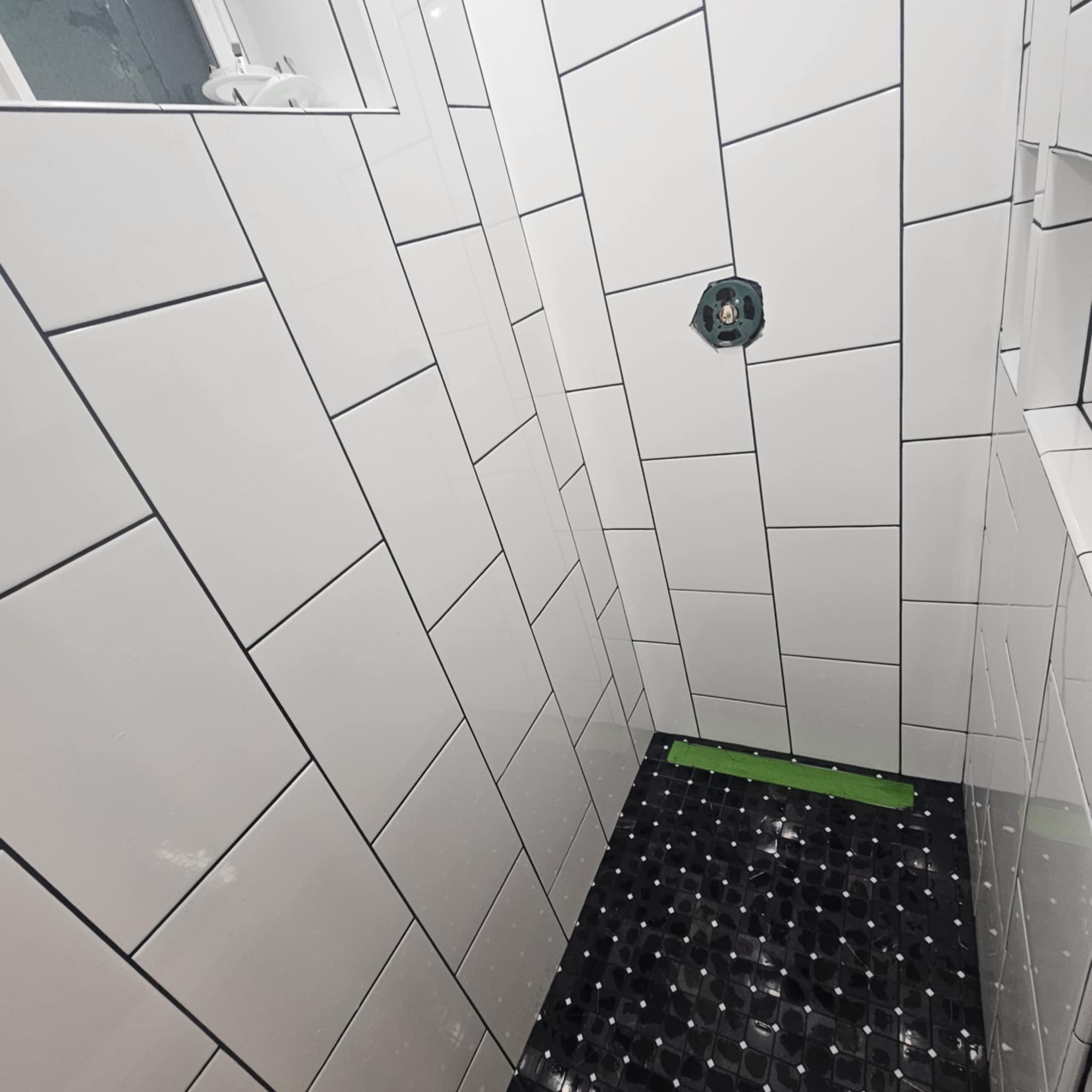 A partially tiled bathroom shower area with white rectangular tiles on the walls and a black hexagon tile floor. A circular hole is cut into the wall, possibly for a fixture to be installed.