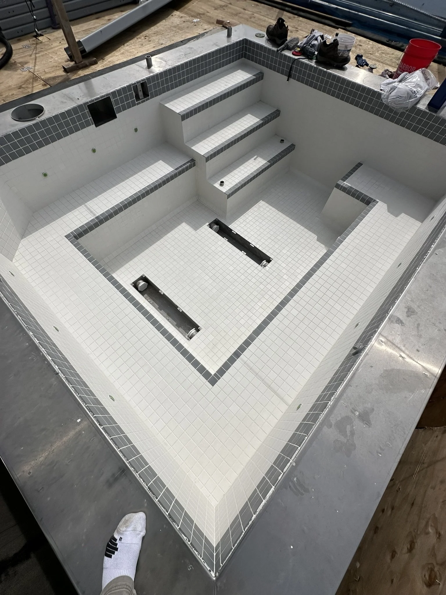 Under construction empty swimming pool with tiled steps and interior, surrounded by construction tools and materials.