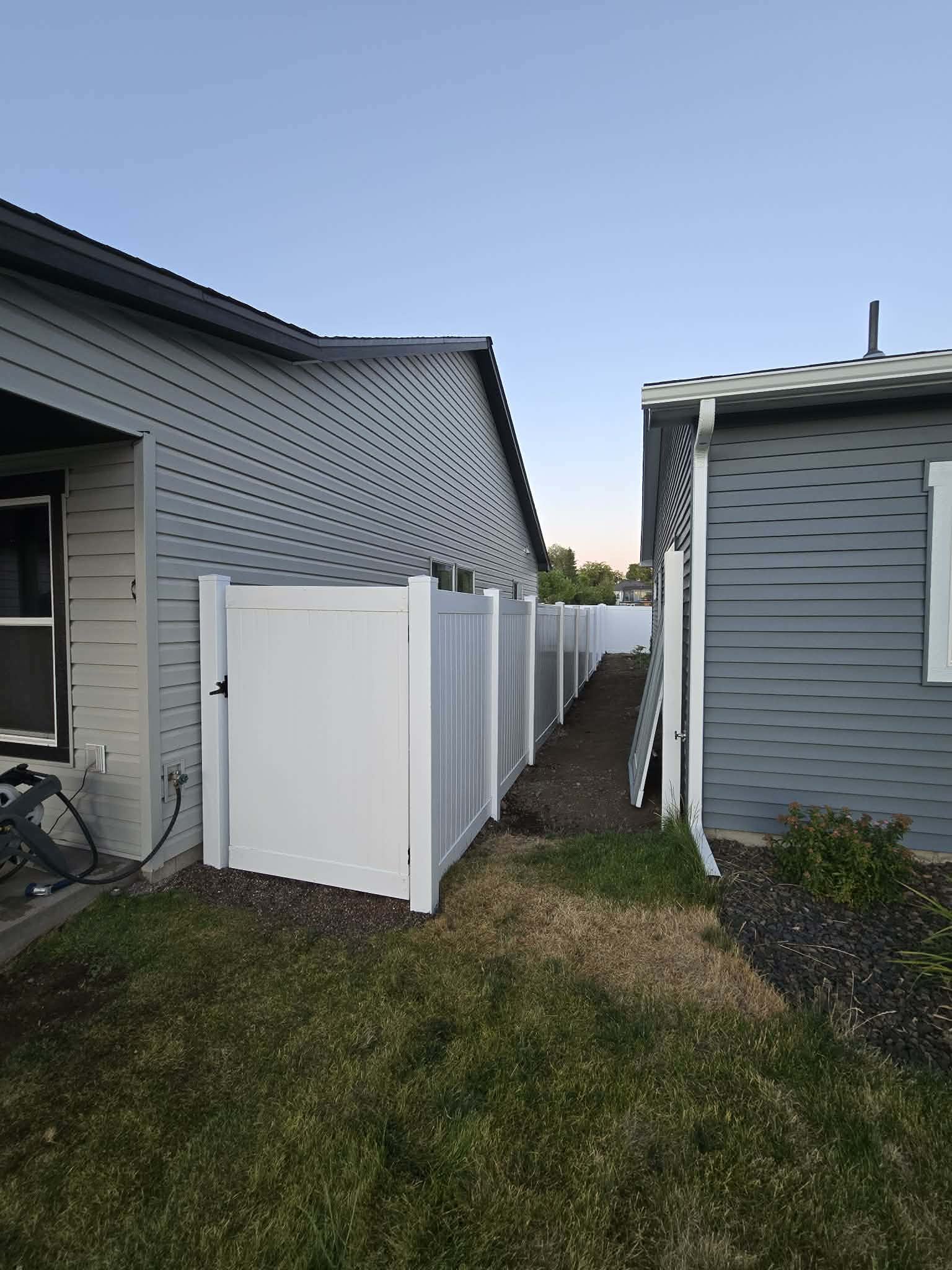 View of a backyard between two houses with a white privacy fence, a small patch of grass, and a narrow dirt pathway.