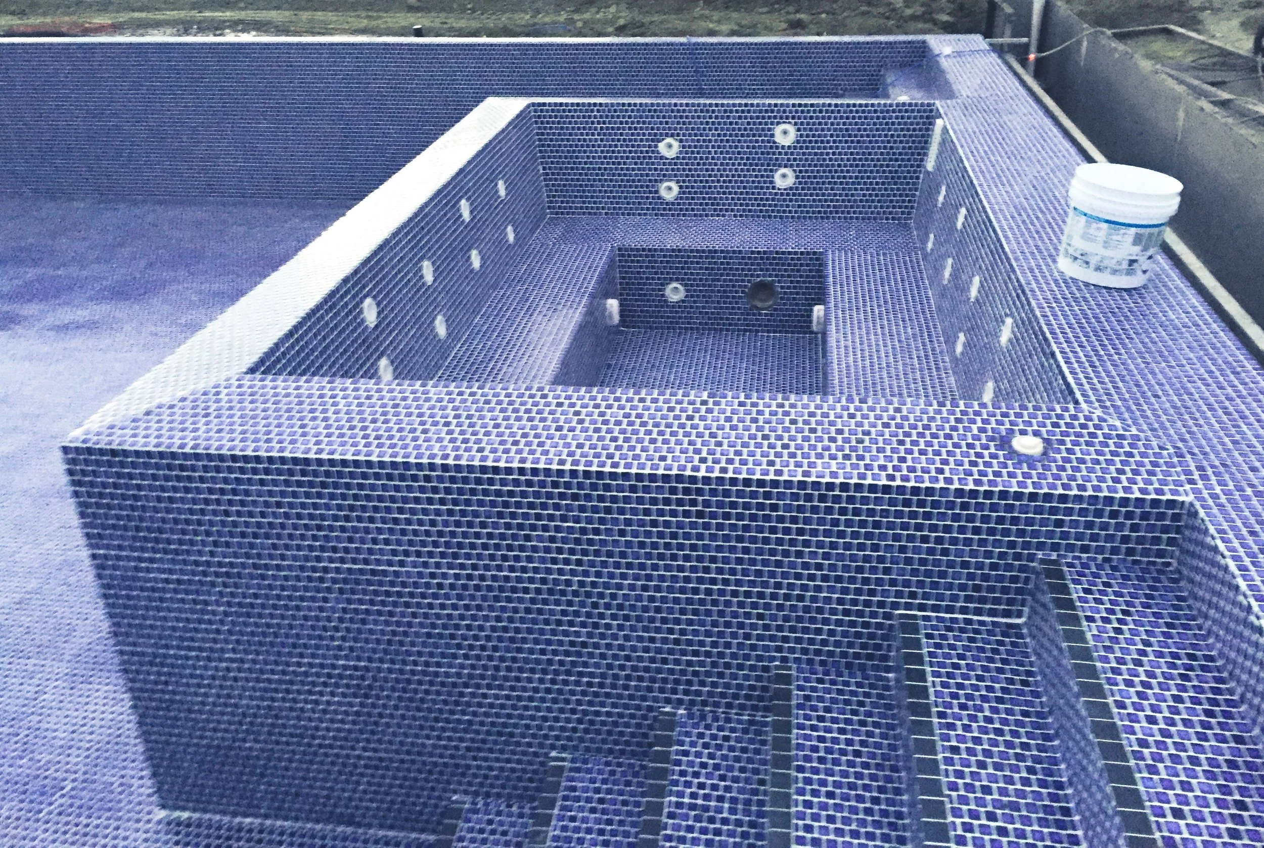 An unfinished hot tub with blue mosaic tiles and a step ladder inside, with a white container on the edge.