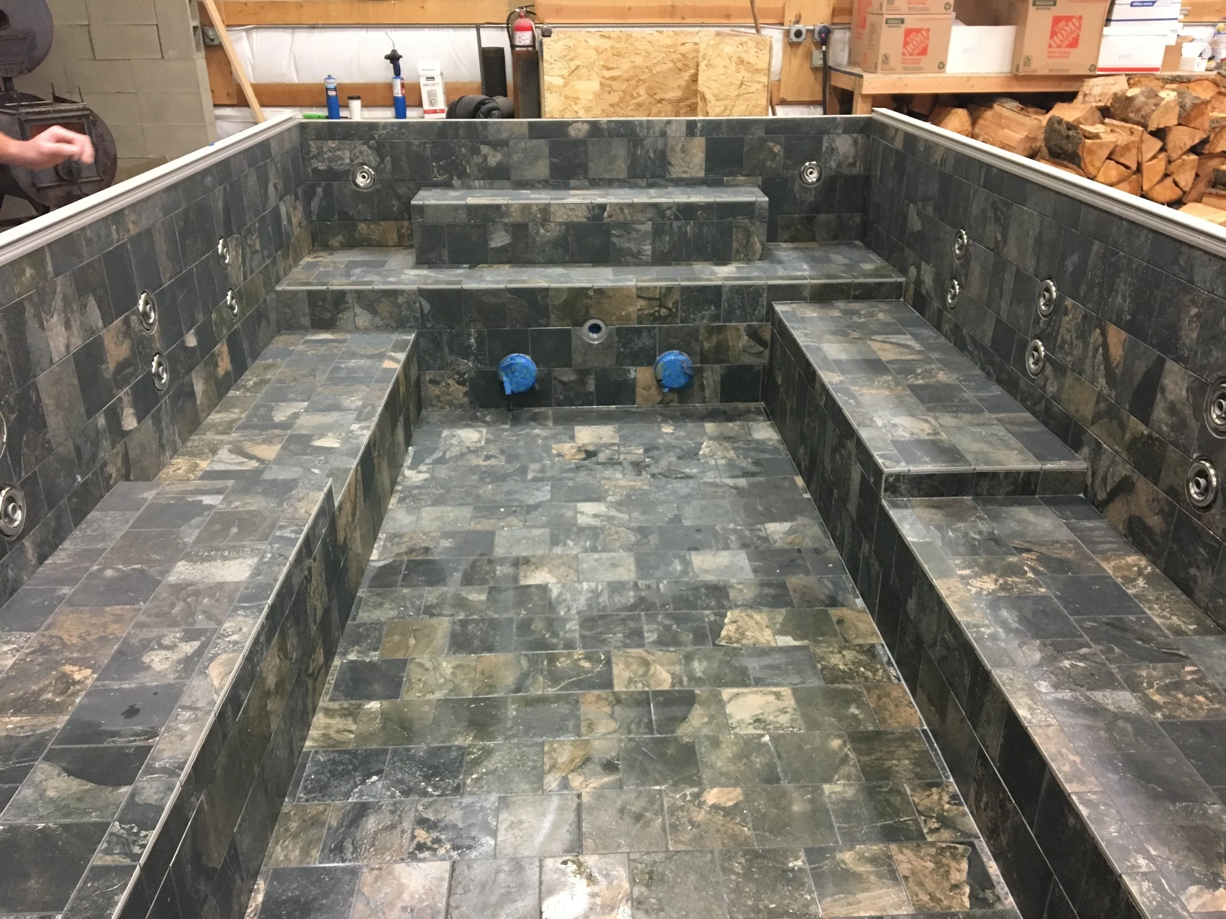 Interior of a tiled hot tub or spa with built-in seating and jets, under construction or renovation, in a workshop or warehouse setting.