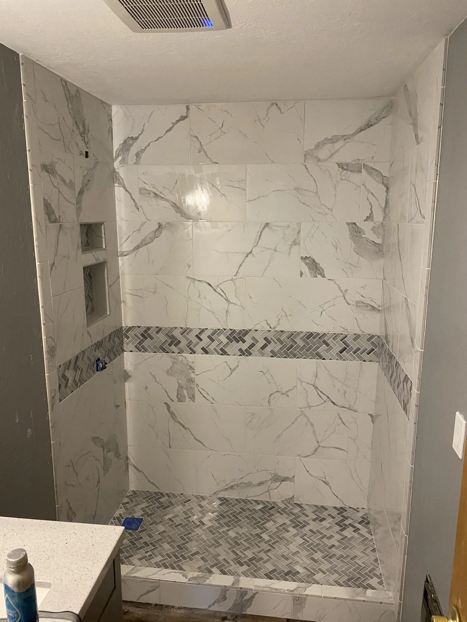 A tiled walk-in shower with large white marble-like tiles with gray veining on the walls, a decorative horizontal strip of small gray herringbone tiles, and a matching herringbone tile floor, with built-in wall niches.
