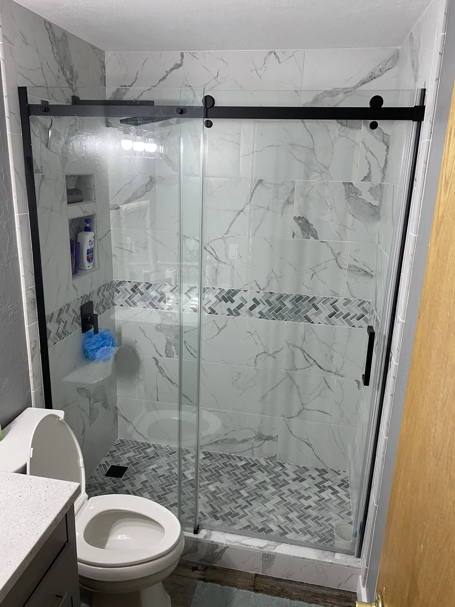 Bathroom with a glass-enclosed shower featuring marble tiles, a built-in niche, and mosaic tile accents on the wall and floor. A toilet is in the foreground and a partial view of a countertop.