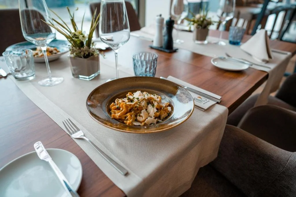 A dining table set with a beige table runner, a plate of pasta with cheese, glasses, and potted plants in a restaurant.