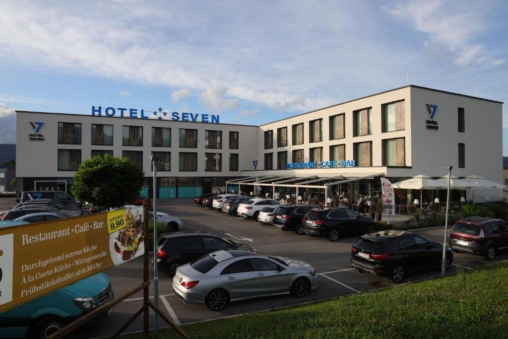 A modern hotel called 'Hotel Seven' with a parking lot in front, outdoor seating at a restaurant with umbrellas, and a sign advertising breakfast buffet for 9.90 euros.