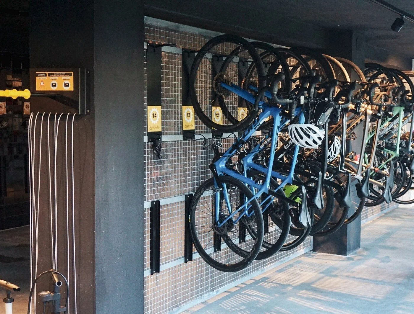 Bicycle parking rack with multiple bikes hanging on black wall mounts, some with helmets, next to a tiled wall section with numbered labels.