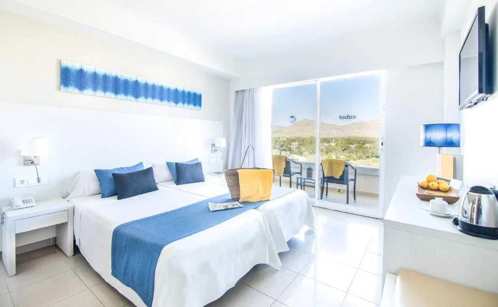 Bright hotel room with a large bed, blue and white bedding, and a balcony with chairs and a view of hills and sky.