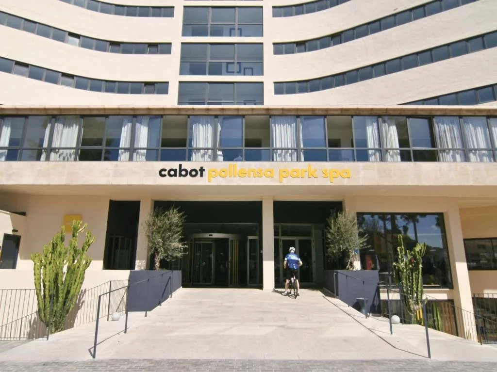 Exterior of the Cabot Pollensa Park Spa hotel with a bicycle rider in front, desert plants on either side, and large windows above.