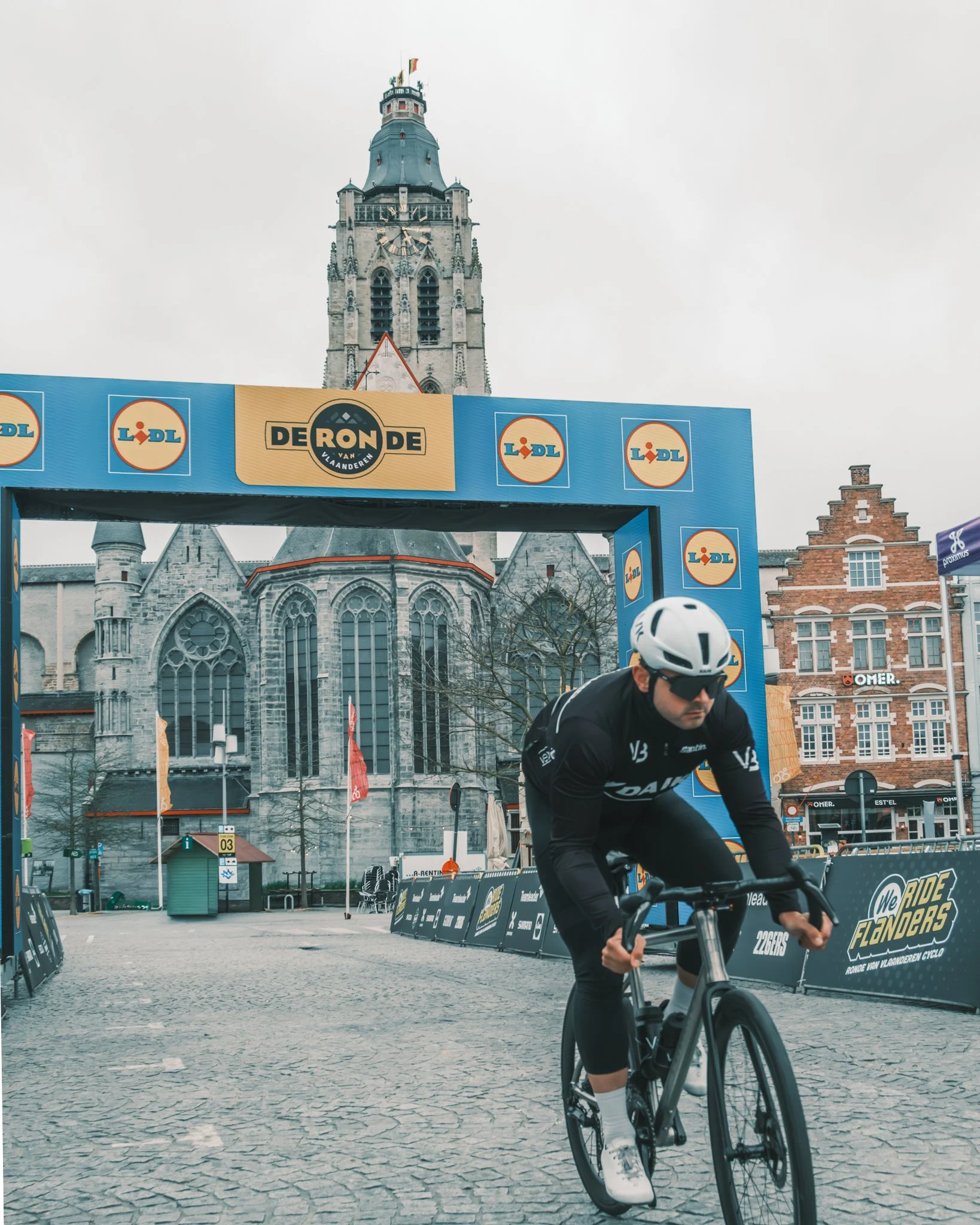 A cyclist wearing black athletic gear riding a black road bike on a cobblestone street with historic buildings and a church in the background. Banners and signs for a cycling event are visible.