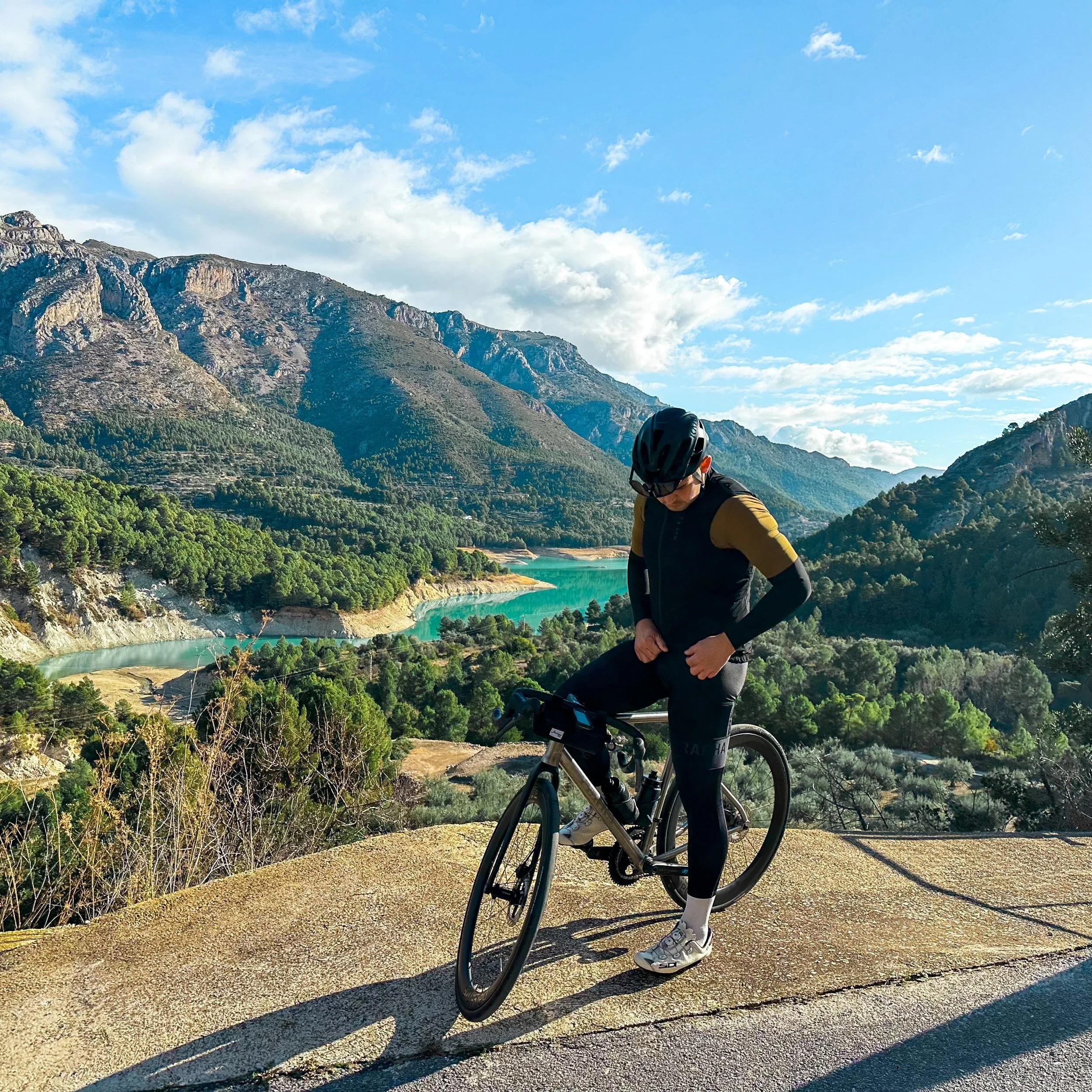 A cyclist wearing a black helmet and yellow and black clothing rides a bicycle on a scenic mountain trail. The background features mountain ranges, green trees, a body of water, and a partly cloudy sky.