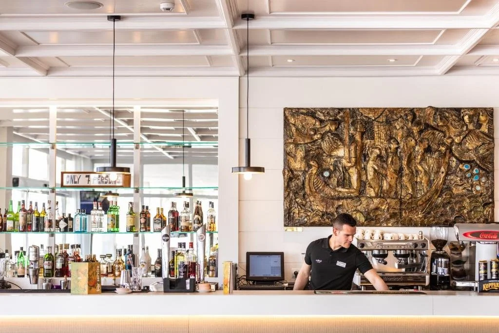 Bar scene with liquor bottles on shelves, a bartender preparing drinks, a coffee machine, and a large artwork on the wall behind the bar.