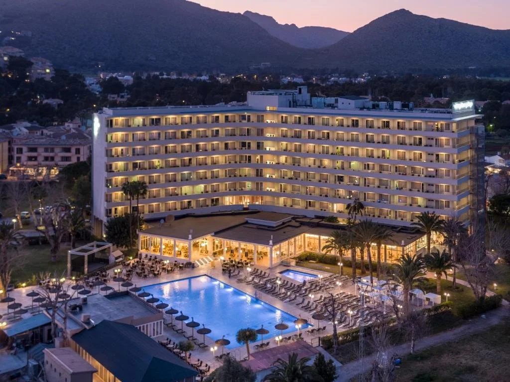 A multi-story hotel with glowing lights at dusk, surrounded by palm trees and an outdoor pool area with chairs and umbrellas.