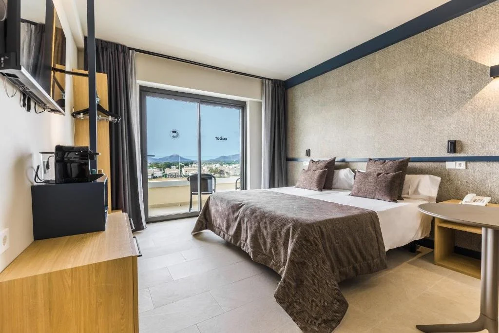 Hotel room with a large bed, two pillows, a bedside table, a sliding glass door with outdoor view, and a balcony with two chairs.