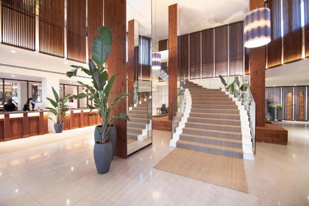 Hotel lobby featuring a staircase with carpeted steps, wooden accents, large potted plants, a reception desk with staff, and modern lighting fixtures.