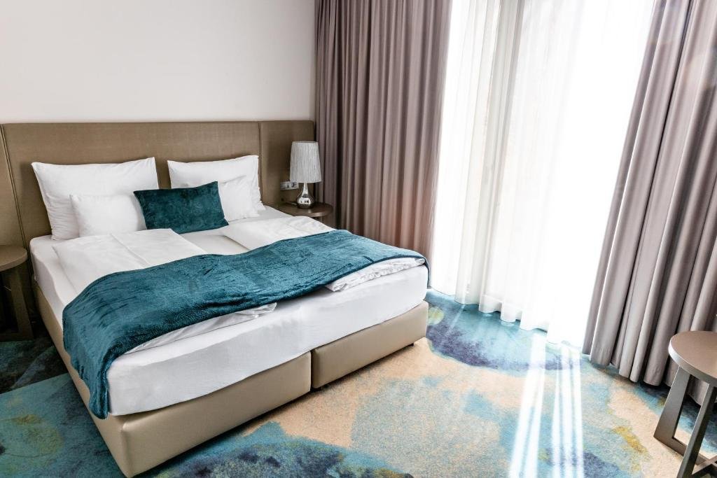 Hotel room with a bed, white linens, teal blanket and pillow, bedside table with lamp, large window with curtains, and colorful carpet.