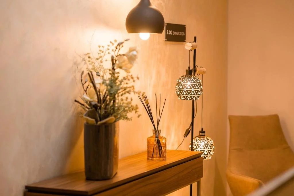 A cozy corner with a wooden side table holding a vase of dried flowers, a reed diffuser, and out-of-focus hanging lamps, with a beige wall and a yellow armchair in the background.