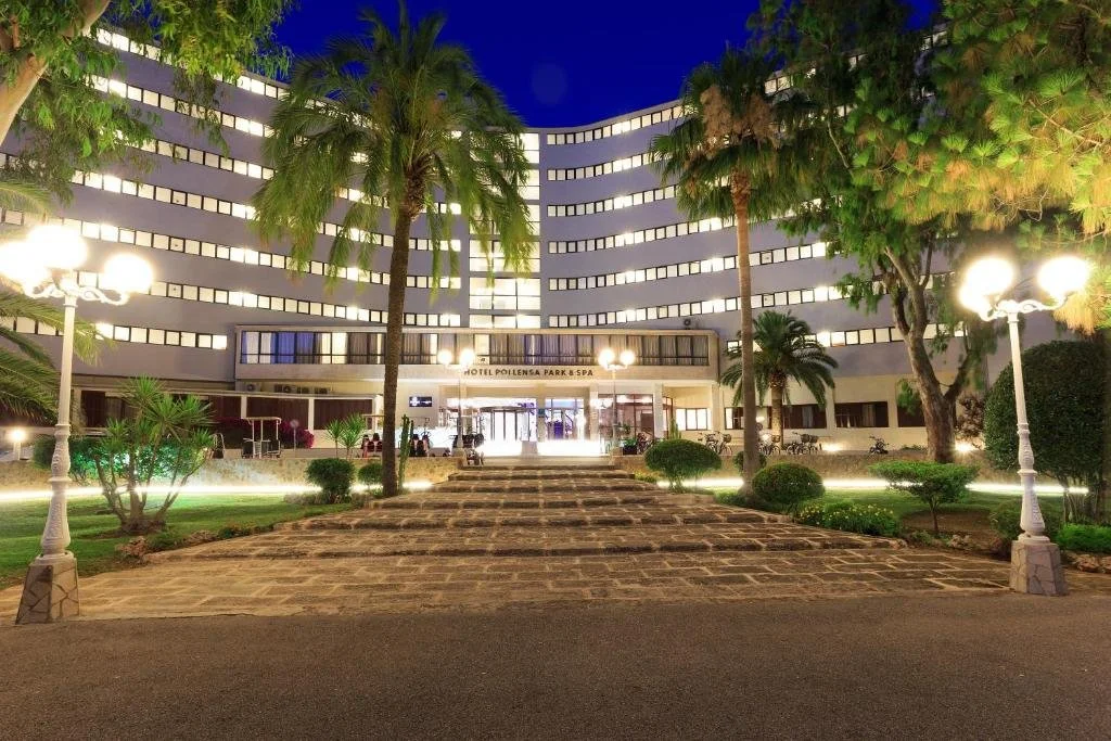 Night view of the hotel Polyhensa Park & Spa with a curved modern white building, palm trees, landscaped garden, and pathway leading to the entrance, illuminated by streetlights.