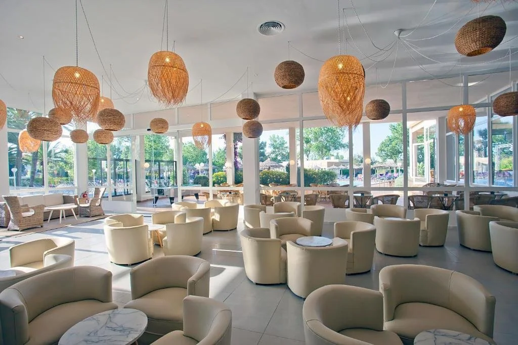 Interior of a modern lounge with cream-colored armchairs, wicker pendant lights hanging from the ceiling, large windows letting in natural light, and tables with marble tops.