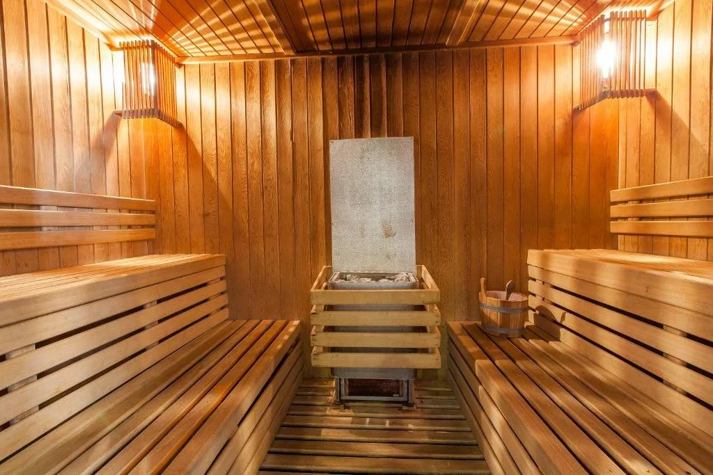 Wood-paneled sauna interior with two benches on each side, a bucket and ladle on the right bench, and a heater with stones at the center.