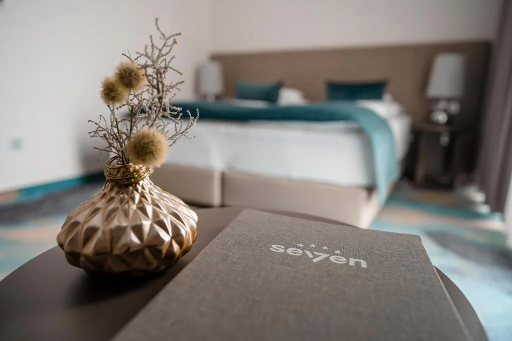 A close-up of a decorative vase with dry flowers placed on a table in a hotel room, with a bed and bedside tables in the background.