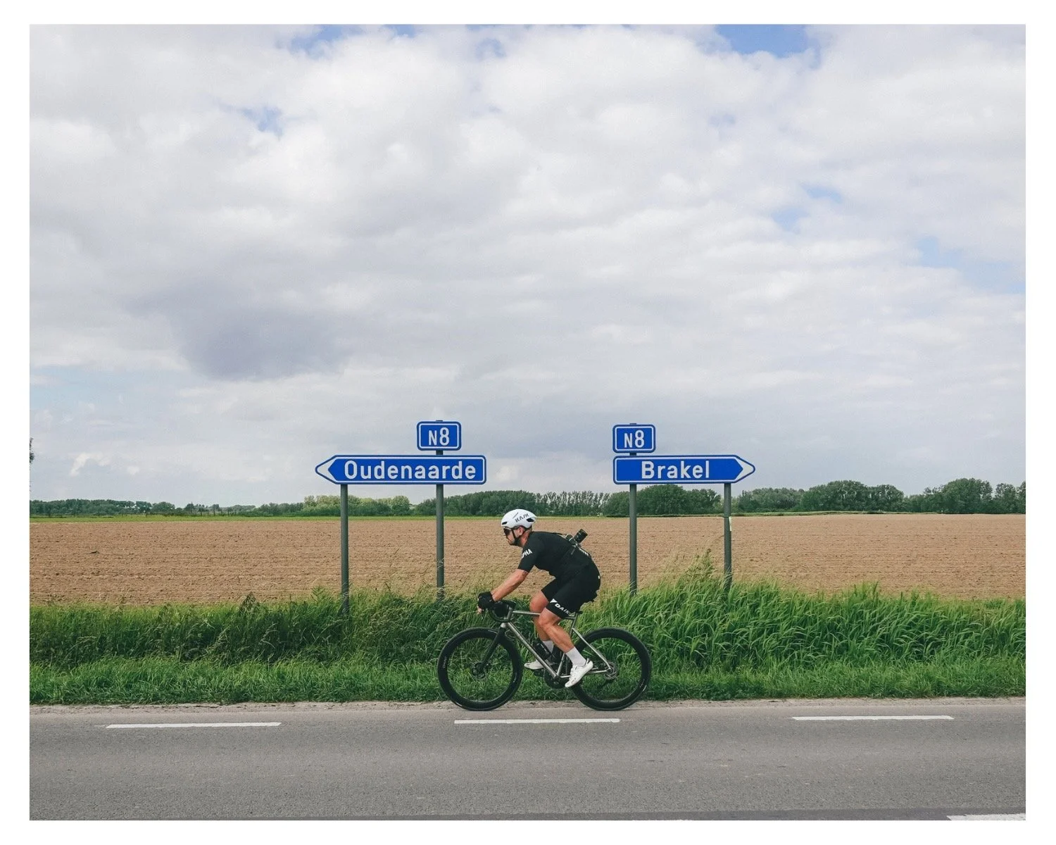 A cyclist wearing a helmet and black attire riding a mountain bike on a rural road with blue directional signs pointing to Oudenarde and Brakel, Belgium, against a backdrop of farmland and cloudy sky.