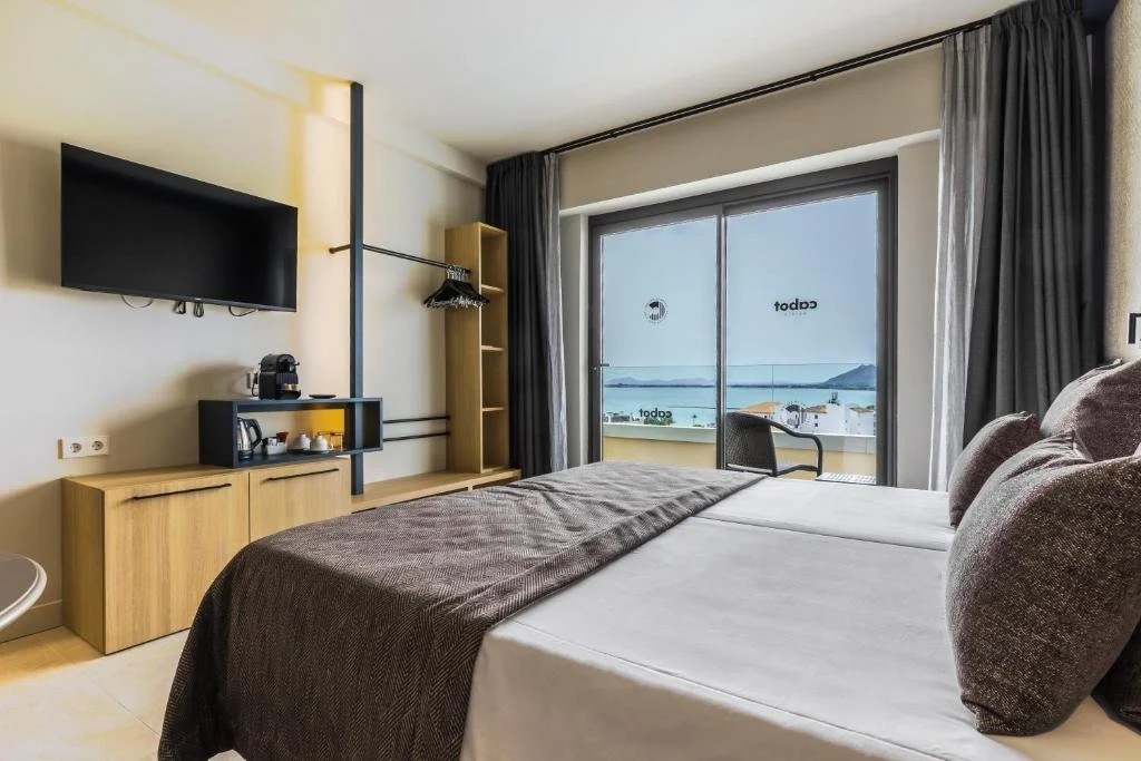 Hotel room with a bed, a wall-mounted TV, a sliding glass door overlooking a balcony with a view of water and hills, dark curtains, and wooden furniture.