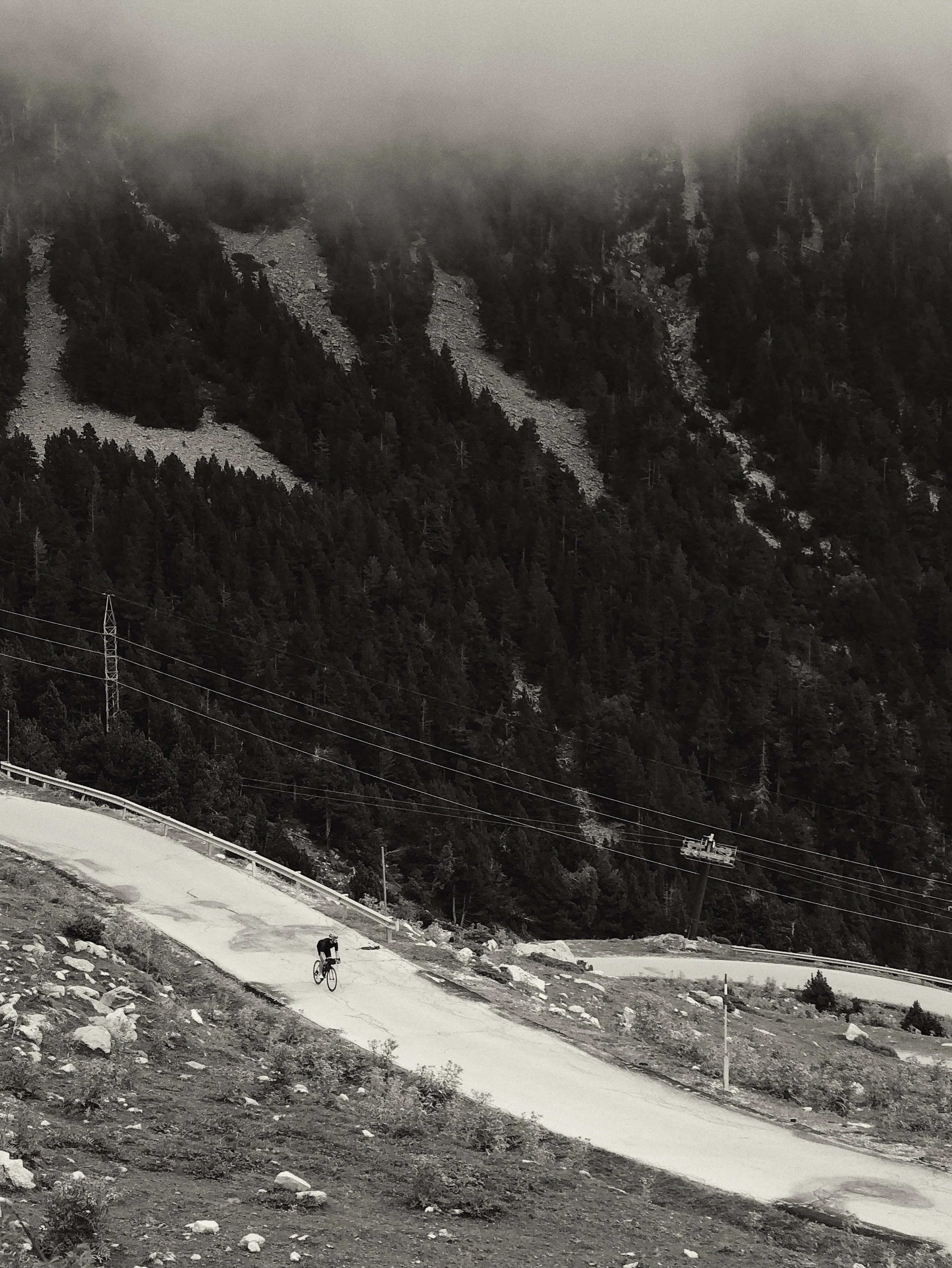 Cyclist riding on a dirt mountain trail with rocky terrain, forested mountain slopes, power lines, and a cable car in the background, under a foggy sky.