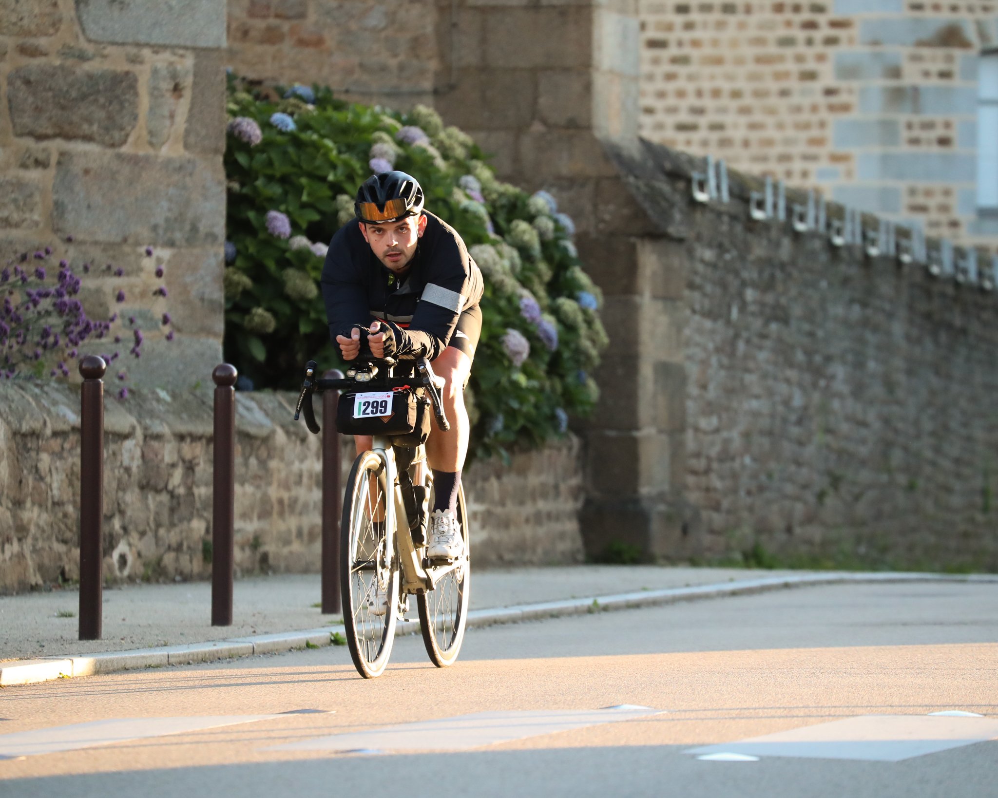 A cyclist riding a bike along a sidewalk in front of a stone wall and flowering bushes, wearing a black helmet and black clothing.