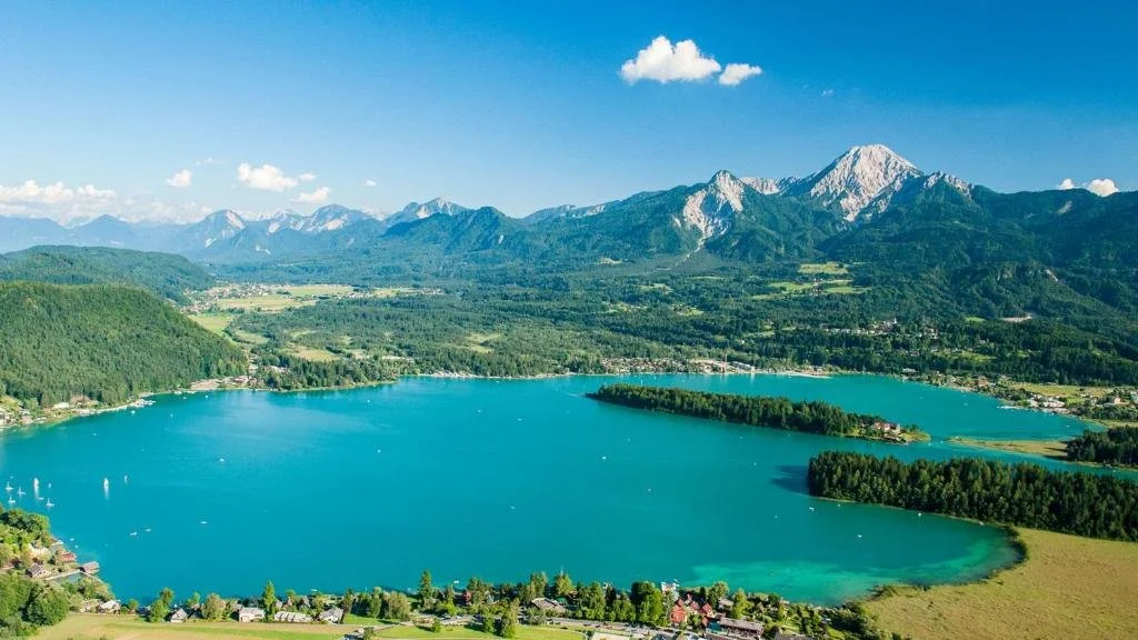 A large blue lake surrounded by green forests and hills, with mountains in the background under a clear sky with a few clouds.