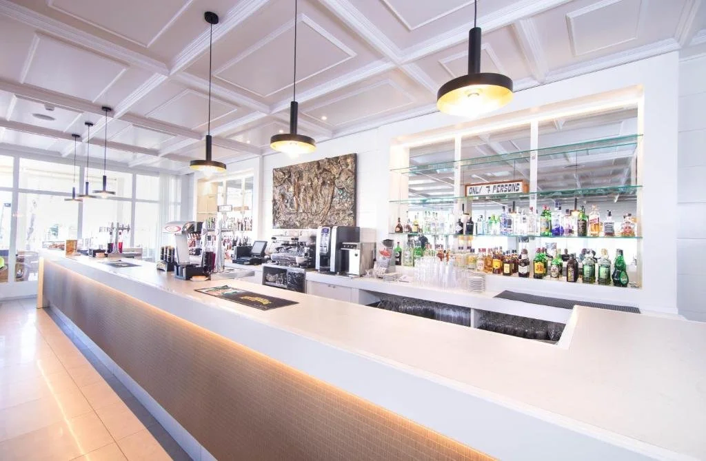 Empty bar counter with various drink dispensers and coffee machines, illuminated by hanging pendant lights, in a bright, modern restaurant or bar with large windows and white decor.