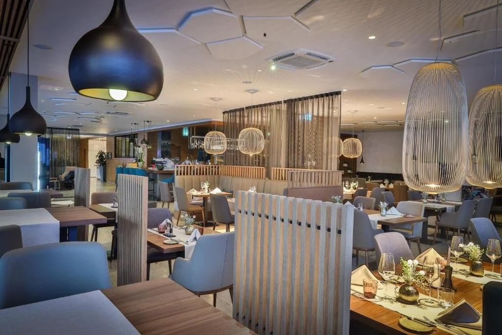 Elegant restaurant interior with various tables set with glassware, napkins, and small flower arrangements. Modern lighting fixtures, including large black and woven pendant lights, illuminate the space with a cozy ambiance.