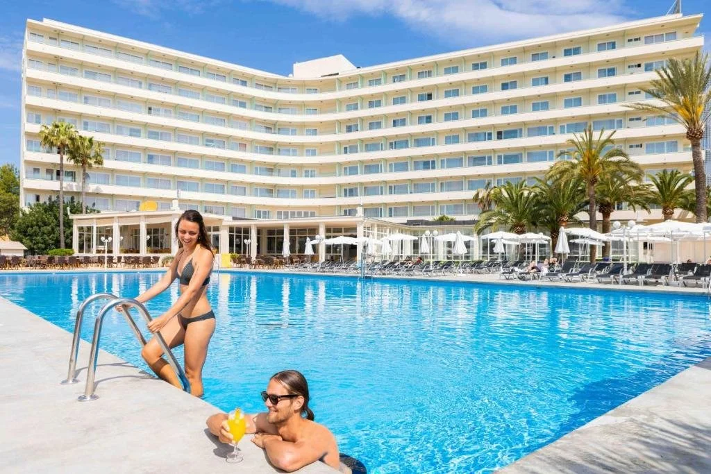A woman in a black bikini is about to enter a large hotel swimming pool, while a man with sunglasses relaxes in the pool with a drink in hand, near a hotel with multiple floors and white balconies.