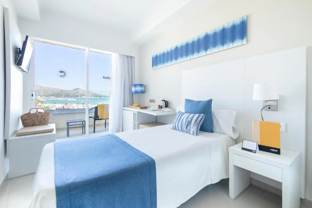 A bright hotel room with a white bed, blue accents, and a view of a harbor through sliding glass doors.