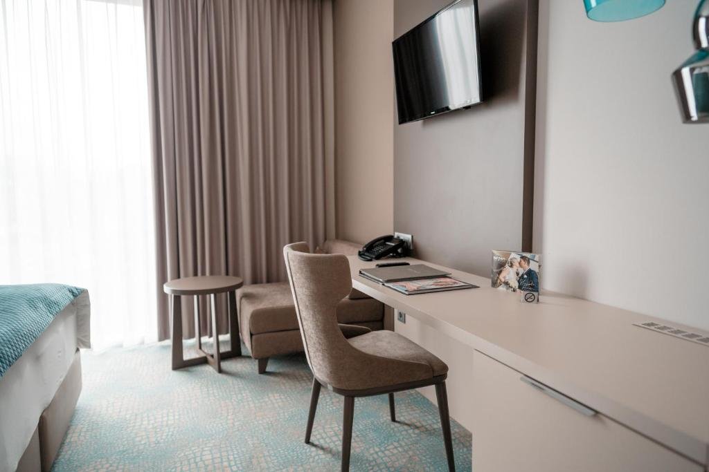 Hotel room with a bed, desk area with a chair, TV, and window with curtains.