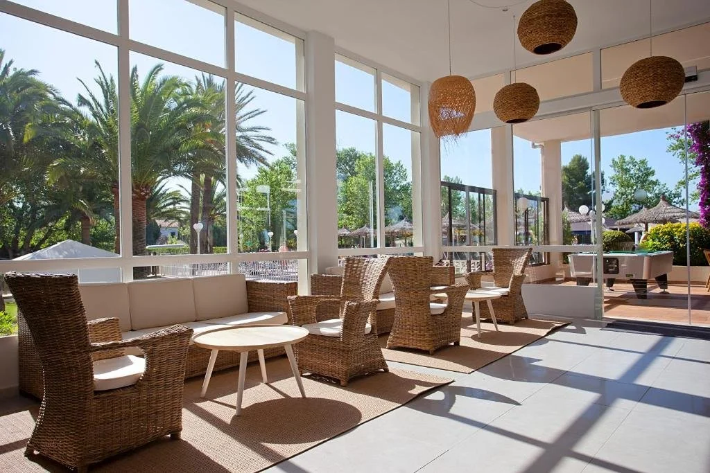 A bright indoor lounge area with wicker chairs, a beige sofa, and round white tables. Large windows and glass doors reveal a sunny outdoor patio with palm trees and umbrellas.