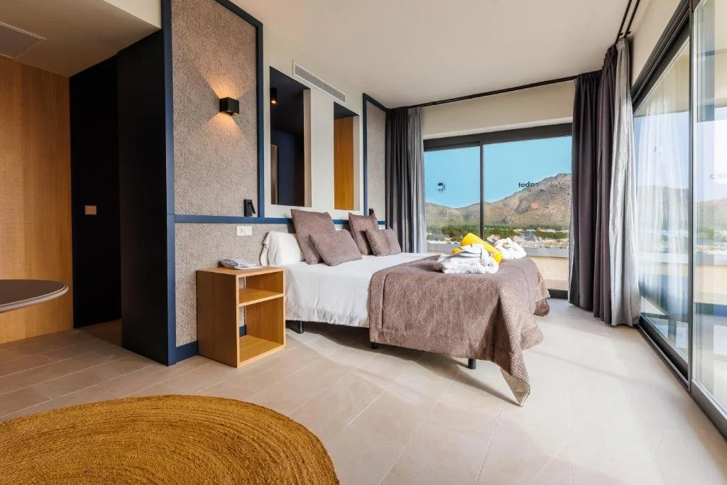 Modern hotel room with large bed, decorative pillows, nightstand, and a folded towel with flowers, large window with scenic mountain view, and sliding glass door leading to balcony.