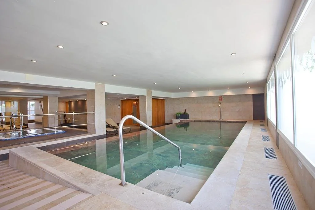 Indoor swimming pool area with stairs, sunlight through large windows, and lounge chairs in the background.