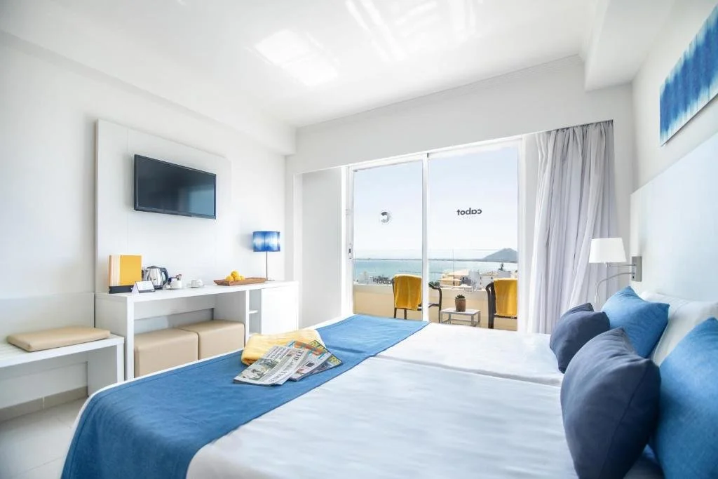 Interior of a hotel room with two beds, a balcony with ocean view, TV, and seating area.