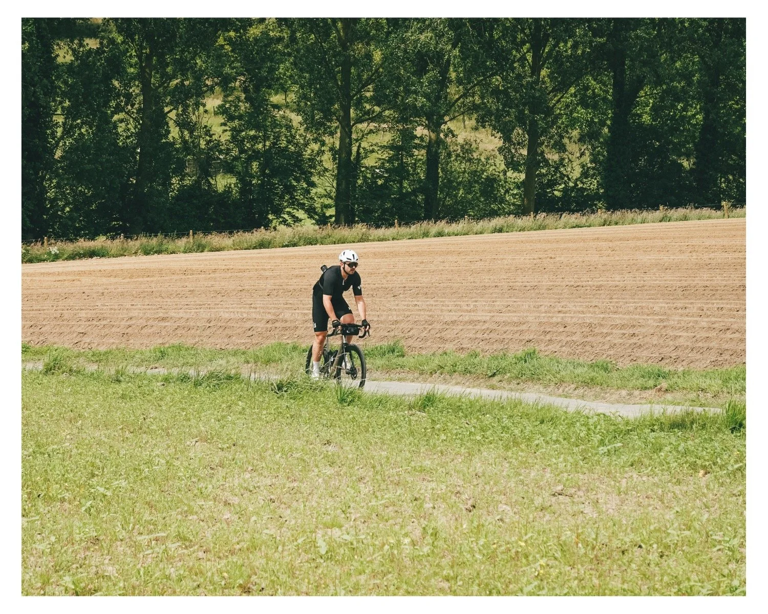 A man riding a bicycle along a dirt path next to a plowed field, with trees in the background on a sunny day.