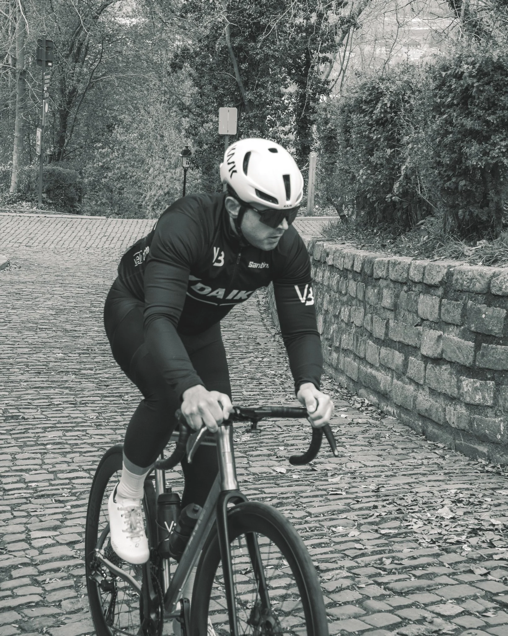 A cyclist wearing a helmet, sunglasses, and athletic clothing riding a bike on a cobblestone path, with trees and foliage in the background.