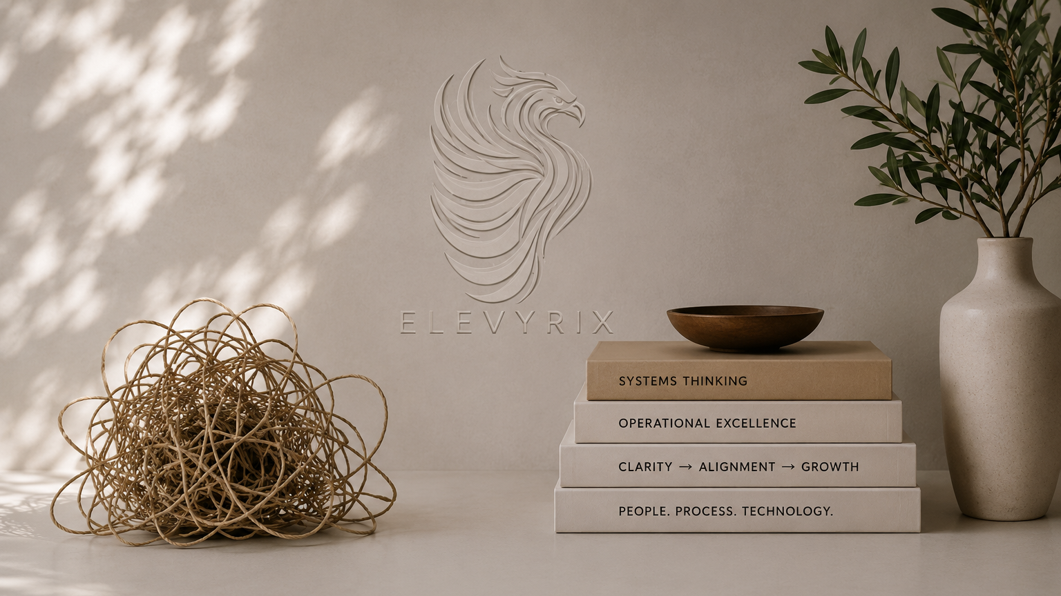A minimalist desk setup with a stack of four books, a bowl on top, a decorative woven ball to the left, and a tall white vase with green leaves to the right. Behind, a logo with a stylized phoenix and the word "ELEVYRIX" is displayed on the wall.