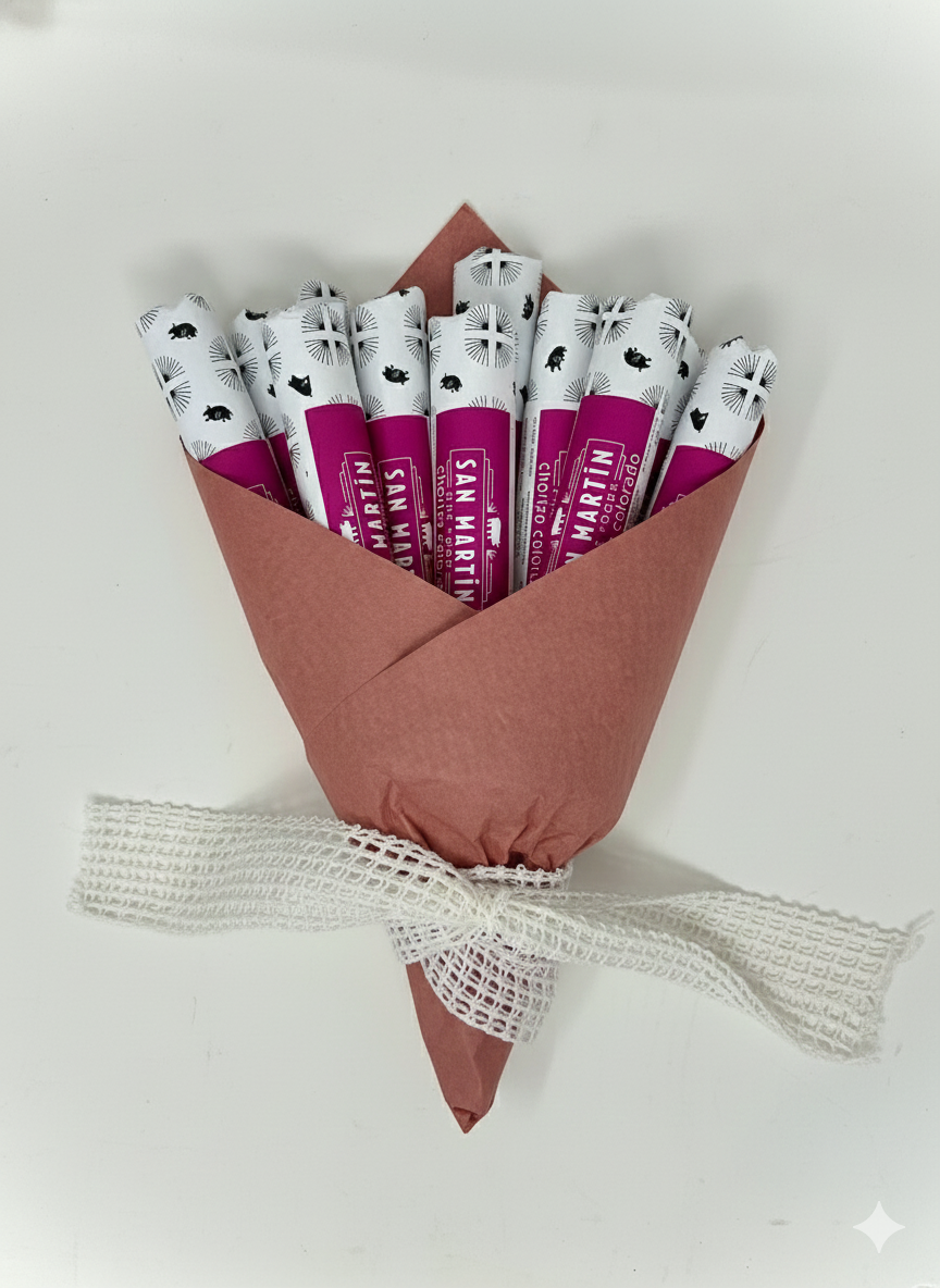 A small bouquet wrapped in pink paper with white and black printed designs, tied with a white mesh ribbon, containing several smoked chorizo sausage links.