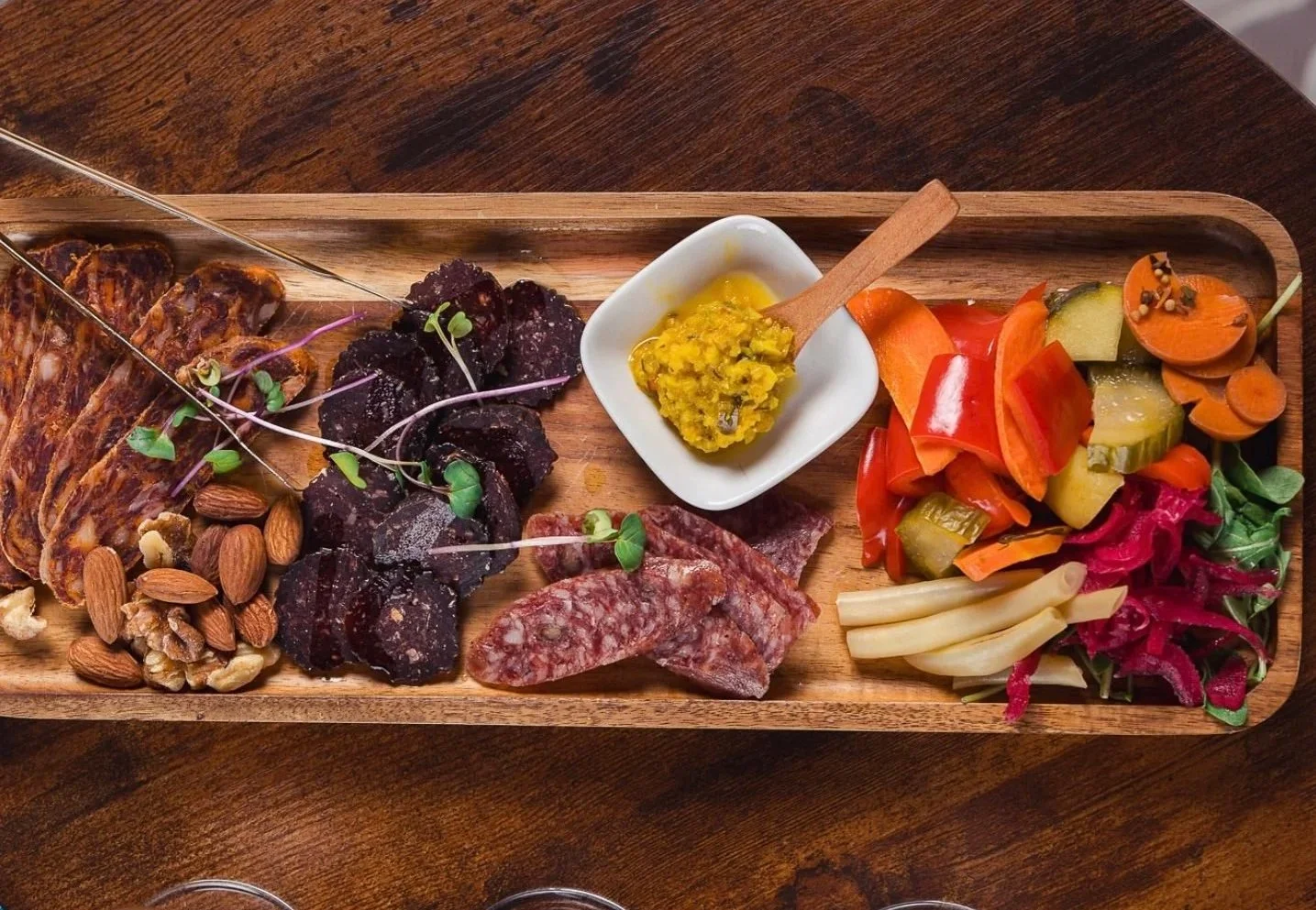 Charcuterie board with sliced meats, nuts, pickled vegetables, and a mustard or relish in a small dish.